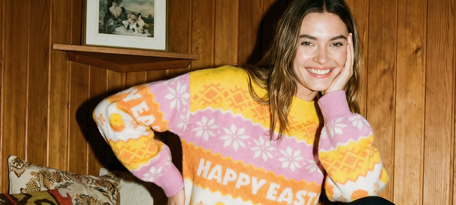 Leende kvinna i färgglad stickad tröja med texten Happy Easter framför en träpanelvägg