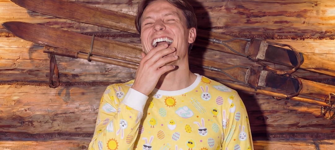 Skrattande man i gul pyjamas med påskmotiv framför vägg med gamla träskidor